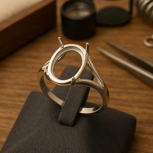 Peut inclure: Un sertissage de bague en argent avec un centre ovale, présenté sur un présentoir noir. La bague comporte des griffes pour maintenir une pierre précieuse. Des outils de joaillerie, dont une pince à épiler et une loupe, sont en arrière-plan.