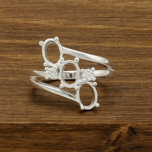 Peut inclure: Une bague en argent avec trois montures ovales et deux petits accents en forme de diamant. La bague a un design unique et ouvert avec une tige fendue. La bague est posée sur un fond en bois.