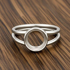 May include: A silver ring with a circular setting and a double band design. The ring is made of a shiny, metallic material and sits on a wooden surface. The ring's design features a central circular setting, perfect for a gemstone or other embellishment.