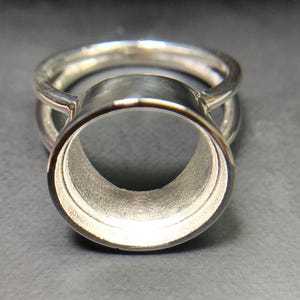 May include: A silver-toned metal ring featuring a prominent circular bezel setting designed for a stone or cabochon. The ring has a smooth, polished finish with a double-band structure that connects to the central circular cup-like setting.
