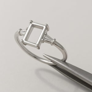 以下が含まれることがあります： 長方形のセンターセッティングとバゲット型のアクセントが付いたシルバーリング。 リングはピンセットで保持されており、その複雑なデザインとカスタマイズの可能性を示しています。 リングのバンドは滑らかで研磨されています。