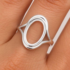 Puede incluir: Un anillo plateado con un diseño ovalado abierto. El anillo está hecho de metal pulido y presenta una estética suave y minimalista. El óvalo es el punto focal, con una banda que se curva alrededor del dedo.