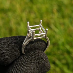 Könnte beinhalten: Eine silberne Ringfassung mit quadratischem Aufsatz, zur Aufnahme eines Edelsteins. Der Ring hat einen schlichten Band und vier Zinken zur Sicherung des Steins. Der Ring wird vor einem schwarzen Hintergrund gehalten.
