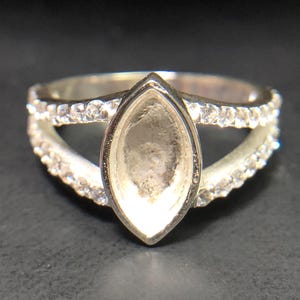 May include: A silver-toned ring featuring a central marquise-shaped empty setting. The band splits into two arms, each encrusted with a row of small, clear, round-cut stones that extend along the sides of the ring, framing the central empty bezel.