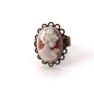 May include: A cameo ring with a white cameo of a woman's profile set in a bronze-colored oval frame with a decorative edge.