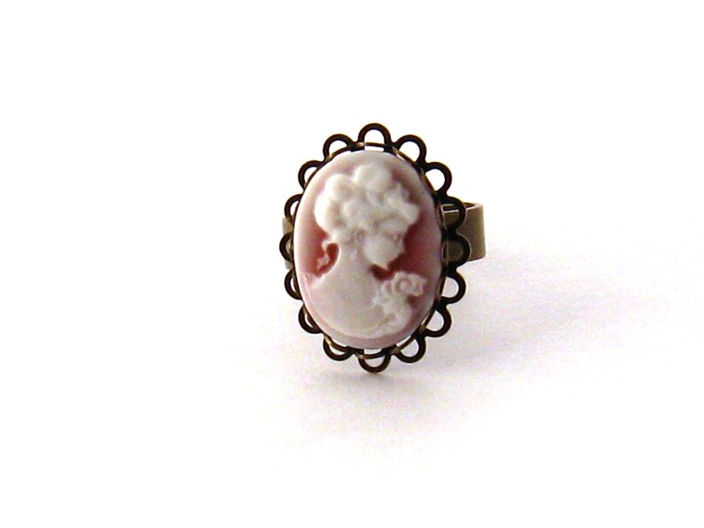 May include: A cameo ring with a white cameo of a woman's profile set in a bronze-colored oval frame with a decorative edge.