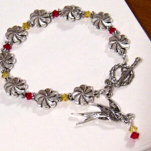 May include: Silver-toned bracelet featuring flower-shaped links and red and yellow crystal accents. A detailed silver bird charm hangs from the bracelet. The bracelet has a toggle clasp.