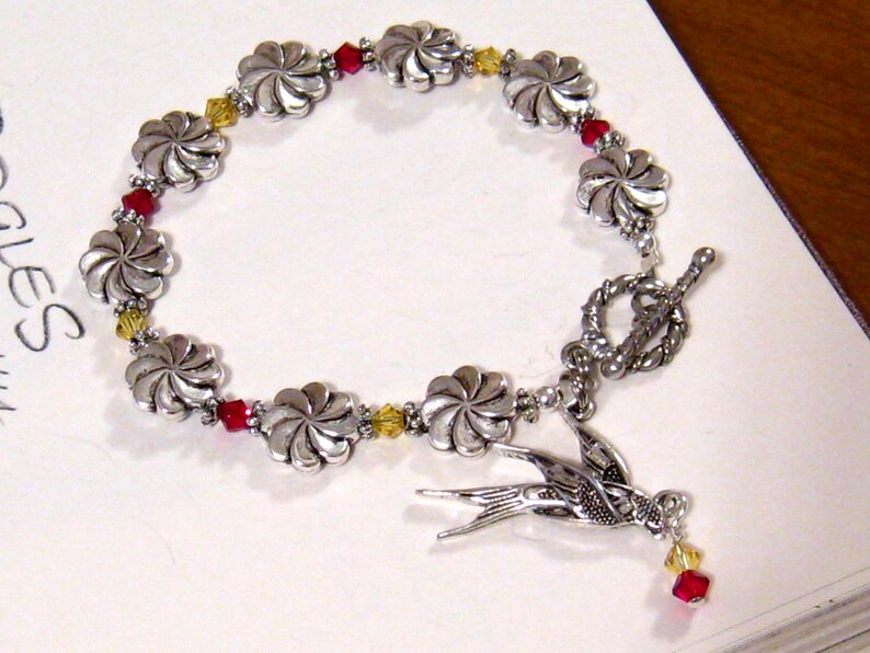 May include: Silver-toned bracelet featuring flower-shaped links and red and yellow crystal accents. A detailed silver bird charm hangs from the bracelet. The bracelet has a toggle clasp.