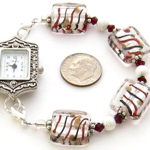 May include: A silver watch with a square face and a white dial. The watch is attached to a bracelet made of clear glass beads with red and black stripes. The bracelet also includes silver beads and red and silver beads.