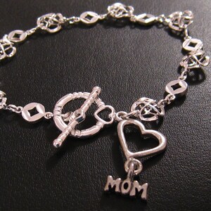 May include: Silver bracelet featuring a heart charm and a 'MOM' charm. The bracelet has a toggle clasp and is made of a delicate chain with small, open heart and square links.