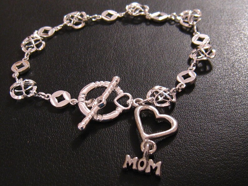 May include: Silver bracelet featuring a heart charm and a 'MOM' charm. The bracelet has a toggle clasp and is made of a delicate chain with small, open heart and square links.