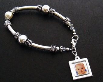 May include: A silver bracelet with a charm featuring a photo of a baby. The bracelet has a pearl bead and silver beads with a textured design.