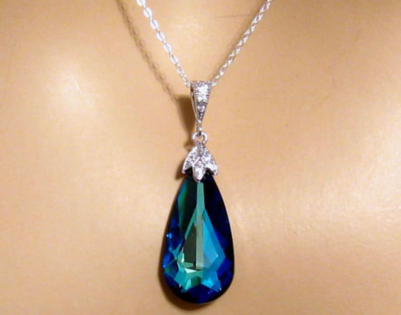 May include: A silver chain necklace with a teardrop-shaped pendant. The pendant is a large, faceted, blue crystal with a green hue.