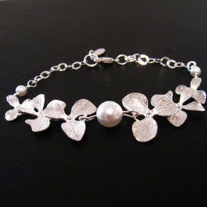 May include: A silver bracelet with a delicate chain and a floral design. The bracelet features a single pearl in the center and is accented with small, white pearls.