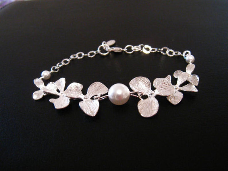 May include: A silver bracelet with a delicate chain and a floral design. The bracelet features a single pearl in the center and is accented with small, white pearls.