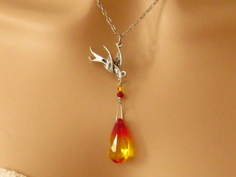 May include: A silver-toned necklace featuring a detailed swallow pendant. The pendant suspends a teardrop-shaped crystal with a gradient of red to yellow. Small red and yellow beads accent the design, creating a striking jewelry piece.