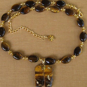 May include: A gold necklace featuring a tiger's eye stone pendant shaped like two elephants facing each other. The necklace is crafted with gold beads and oval-shaped tiger's eye stones.
