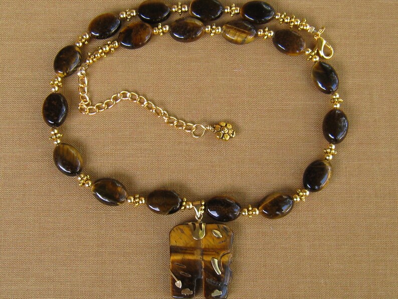 May include: A gold necklace featuring a tiger's eye stone pendant shaped like two elephants facing each other. The necklace is crafted with gold beads and oval-shaped tiger's eye stones.