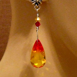 May include: A single earring with a silver-tone floral stud. The earring features a wire with red and yellow crystals, and a large, teardrop-shaped, faceted crystal that transitions from yellow to red.