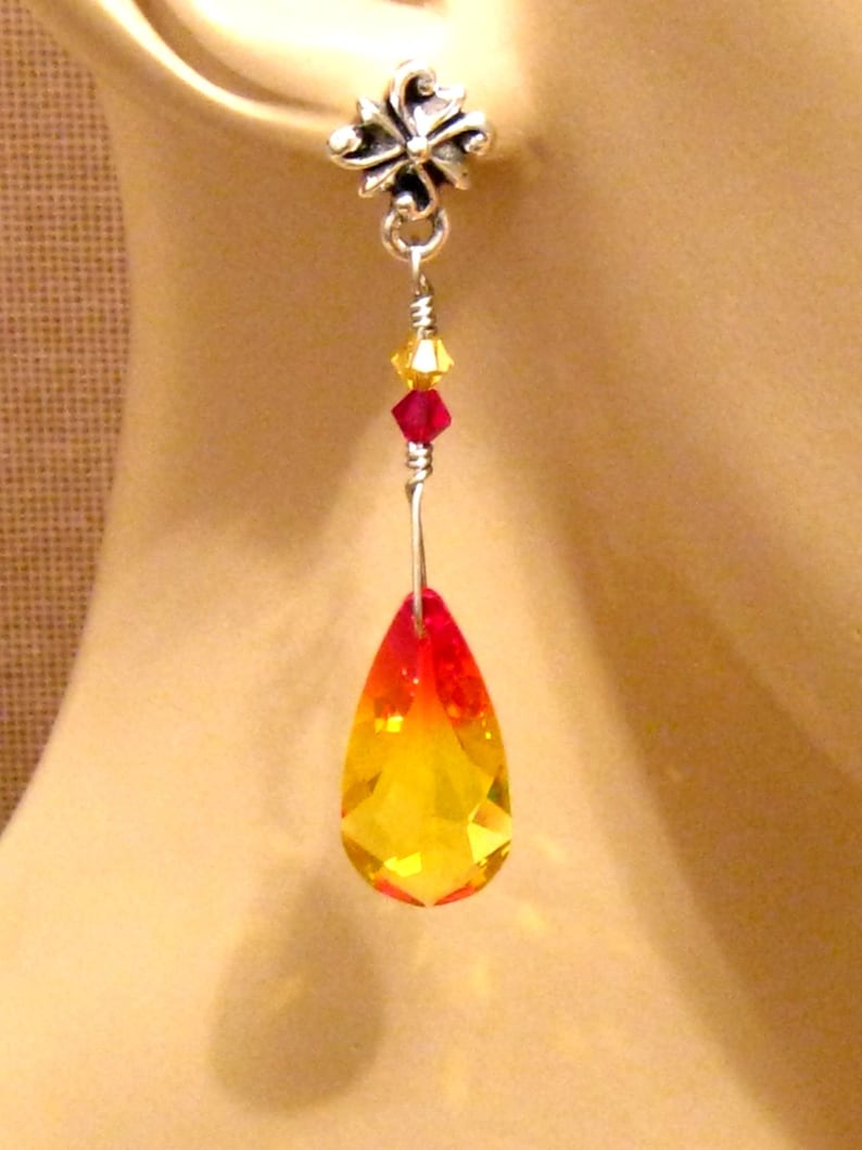 May include: A single earring with a silver-colored floral stud. A wire descends, holding yellow and red crystals, leading to a large, teardrop-shaped crystal that transitions from red to yellow. The earring is designed to dangle.