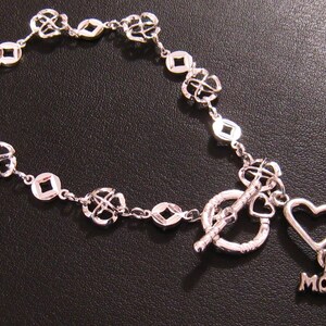 May include: A silver bracelet with a toggle clasp and a heart charm with the word "MOM" on it. The bracelet is made of a chain with small, open squares and four-leaf clover designs.