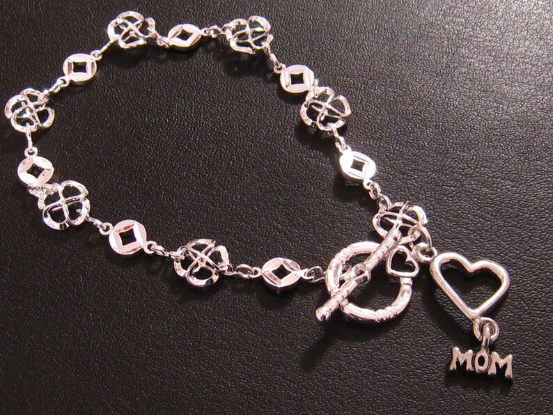 May include: A silver bracelet with a toggle clasp and a heart charm with the word "MOM" on it. The bracelet is made of a chain with small, open squares and four-leaf clover designs.