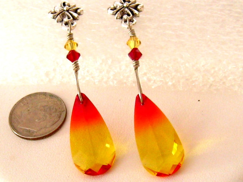 May include: A pair of dangle earrings featuring teardrop-shaped, faceted crystals that transition from red to yellow. The earrings have silver-tone metal accents, including floral studs and wire detailing, with red and yellow beads.