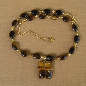 May include: A gold chain necklace with a tiger's eye stone pendant in the shape of two elephants. The necklace is made with brown tiger's eye beads and gold beads.