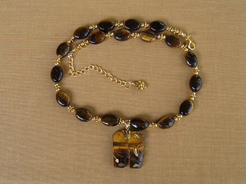 May include: A gold chain necklace with a tiger's eye stone pendant in the shape of two elephants. The necklace is made with brown tiger's eye beads and gold beads.