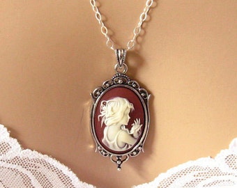 Op de afbeelding: Zilverkleurige ketting met een ovale hanger. De hanger toont een crèmekleurige cameo van een vrouw tegen een bordeauxrode achtergrond, gevat in een sierlijke zilveren rand. De ketting heeft een fijne zilveren schakel.