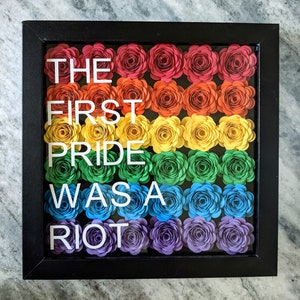 May include: A framed artwork with a rainbow of paper flowers and the text "THE FIRST PRIDE WAS A RIOT" in white letters.