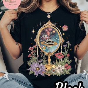 May include: Black t-shirt featuring a celestial design within a gold-framed mirror, surrounded by colorful flowers. The mirror reflects a planet with a striped pattern. The word "Black" is written in white script at the bottom.