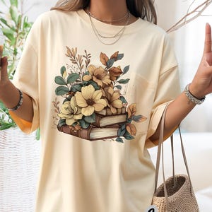 Puede incluir: Camiseta de color marfil con un diseño floral de flores amarillas, hojas verdes y libros marrones. La camiseta tiene un bolsillo y la palabra "Ivory" en fuente cursiva. La camiseta es de corte holgado.