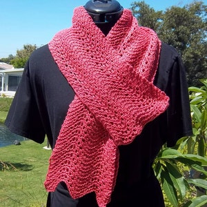 May include: A coral-colored, open-weave scarf is draped over a black form. The scarf has a textured appearance with a scalloped edge. The scarf is made of a soft, lightweight material, perfect for adding a touch of color and warmth.