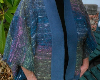 May include: A woman wearing a blue and green woven jacket with a blue collar and a black shirt. She is standing in front of a wooden bench.