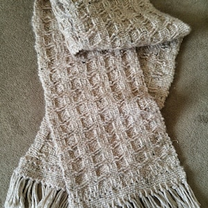 Peut inclure: Une écharpe en tricot beige avec un motif tissé et des franges aux extrémités.