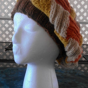 Può includere: Un cappello lavorato a maglia con un motivo a righe colorate in tonalità di marrone, arancione, giallo e beige. Il cappello è esposto su una testa di manichino bianca.