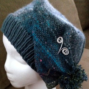 May include: A blue and green knitted hat with a pom-pom and a silver wire accent. The hat is on a white mannequin head.