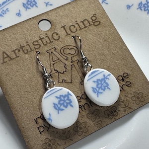 May include: White and blue floral design earrings with silver hooks. The earrings are oval-shaped and feature a delicate blue floral pattern on a white background. The earrings are displayed on a brown card with the text "Artistic Icing".