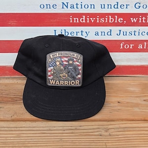 May include: A black baseball cap featuring a rectangular patch on the front. The patch displays an American flag background with two uniformed figures and a German Shepherd dog. The text on the patch reads MY PRONOUN IS WARRIOR.