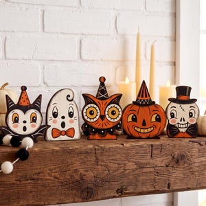 May include: A set of five wooden Halloween decorations featuring a bat, a ghost with a bow tie, an owl in a party hat, a jack-o'-lantern, and a skull wearing a top hat. Each figure is painted in orange, black, and white with a vintage aesthetic.