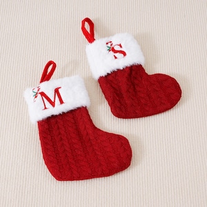 Peut inclure: Deux bas de Noël rouges avec une bordure en fausse fourrure blanche et des boucles de suspension rouges. Les bas ont un motif en tricot torsadé. L'un des bas porte la lettre "M" brodée en rouge, l'autre la lettre "S".