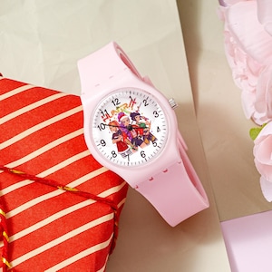 May include: A light pink analog wristwatch with a silicone strap and a white dial featuring black numerals and a colorful graphic illustration of three characters with the text HUNTER in red and green letters above the central character figures.