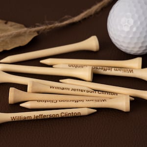 Op de afbeelding: Een verzameling lichtgekleurde houten golftees, elk gegraveerd met de naam "William Jefferson Clinton". Twee witte golfballen zijn zichtbaar op de achtergrond. De tees liggen op een bruin oppervlak.
