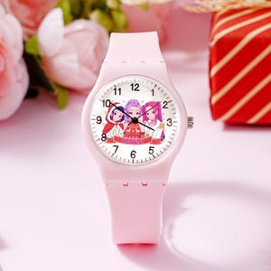 May include: A light pink analog wristwatch with a silicone strap and a white face. The dial features black numerals and a central graphic illustration of three animated characters wearing holiday-themed clothing with the text "HOLIDAY X" below.