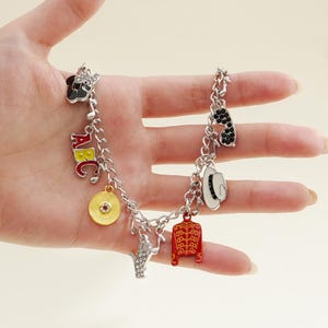 Op de afbeelding: Zilveren bedelarmband met muziekthema-bedels. Bedels zijn onder meer een zwarte strik, zonnebril, een witte hoed, een rode jas, een gouden plaat en de letters ABC. De armband wordt in een hand gehouden.