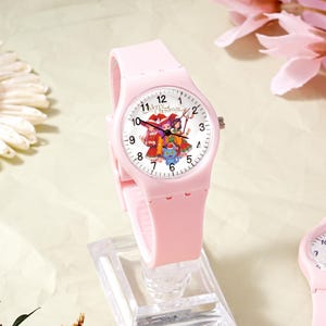 May include: A light pink analog wristwatch with a silicone strap and a white dial. The dial features black numerals, black hands, and a colorful illustration of three anime-style characters wearing Santa hats with the text Merry Christmas.