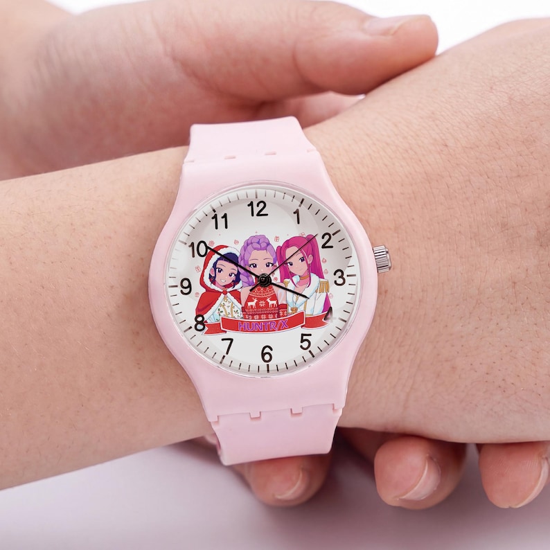 May include: A light pink analog wristwatch with a white face featuring three illustrated characters in festive attire. The watch has black hour, minute, and second hands, black numerical hour markers, and a smooth, solid light pink silicone strap.