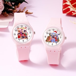 May include: Two light pink analog wristwatches with silicone bands. The left watch face features three cartoon characters and the text "HUNTRX". The right watch face displays a blue cat character wearing a Santa hat with other figures.