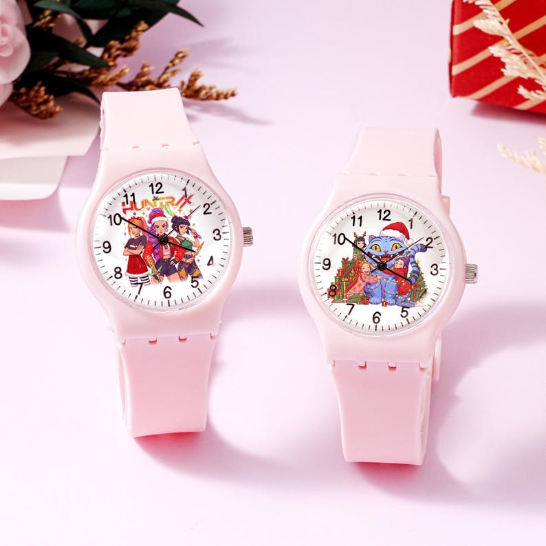 May include: Two light pink analog wristwatches with silicone bands. The left watch face features three cartoon characters and the text "HUNTRX". The right watch face displays a blue cat character wearing a Santa hat with other figures.